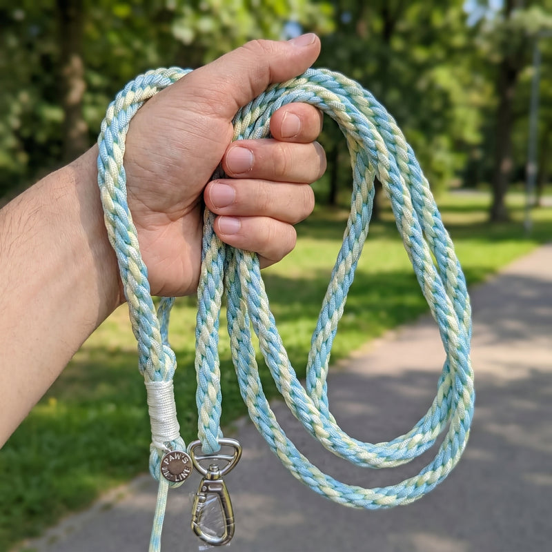 A close-up of a person firmly gripping a coiled Mint Cream Traverse Braided Paracord Leash during an outdoor walk. color:all